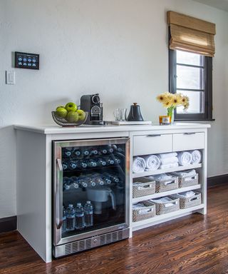 Section of home gym with drinks refrigerator and storage for towels on shelving, coffee machine and fruit bowl with apples above