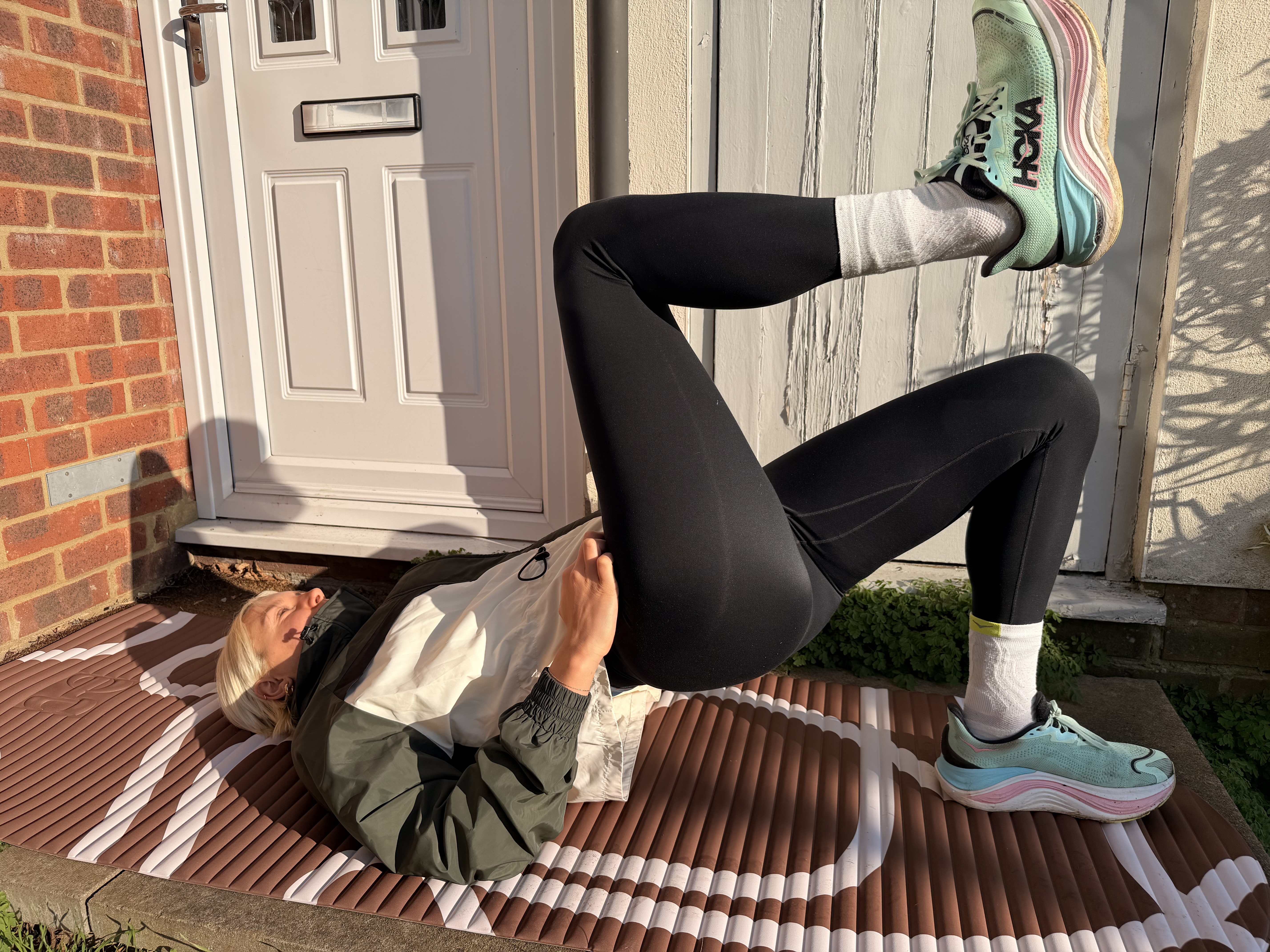 A woman in running clothing lying on her back on a yoga mat with her hips raised off the floor and one leg in the air