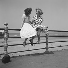 Two women chatting on the railings in Blackpool. The picture was taken by Bert Hardy using a single-focus box camera. The women are former Tiller Girls Pat Wilson (right) of Bridgend, mid Glamorgan, and Wendy Clarke of Southall in London. Blackpool Railings, 1951. Images provided by Getty Images Archive, home of the Picture Post collection, in support of Bert Hardy: Photojournalism in War and Peace. Credit: Bert Hardy via Getty