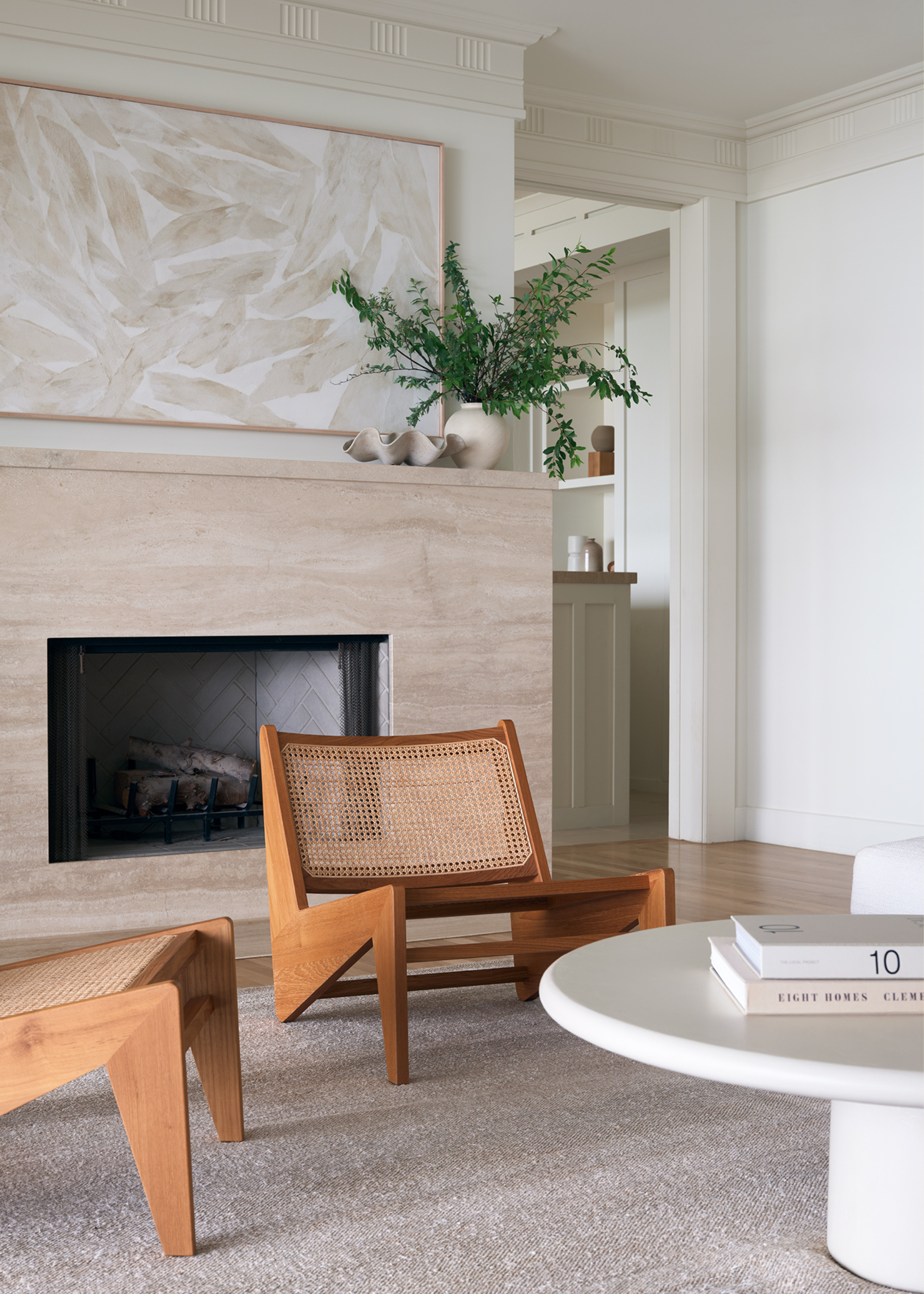 A living room with white walls, a framed minimalist painting, a vase of stems and aceramic bowl on the mantel of a stone fireplace by a pair of rattan wood chairs and a round white coffee table with books on a beige area rug