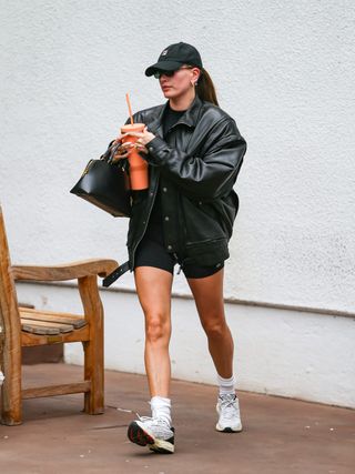 Hailey Bieber wearing a black leather jacket, black t-shirt, black biker shorts, sneakers, and baseball cap