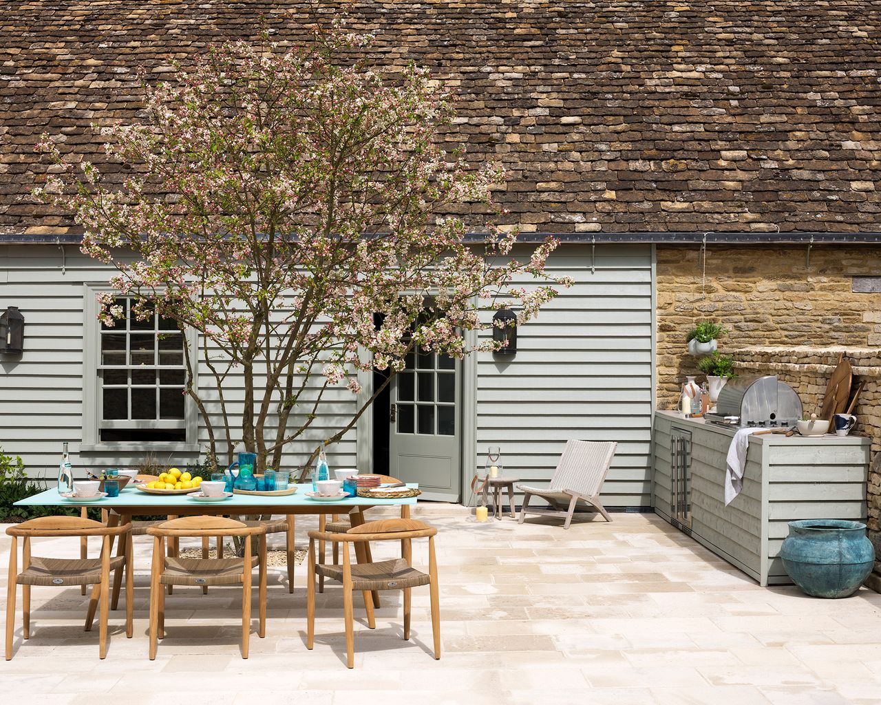 How to plan an outdoor kitchen in a traditional pale gray home&#039;s patio area, with wooden table and chairs and pale stone pavers.