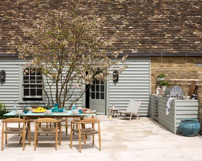 How to plan an outdoor kitchen in a traditional pale gray home's patio area, with wooden table and chairs and pale stone pavers.