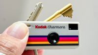 A hand holds a novelty keychain that resembles a Kodak camera, featuring the name "Kodak Charmera" and colorful stripes