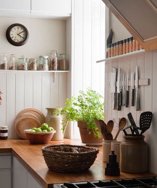 A rustic style kitchen, with plants on the window sill, knifes hanging on the wall, and a woven basket on the counter