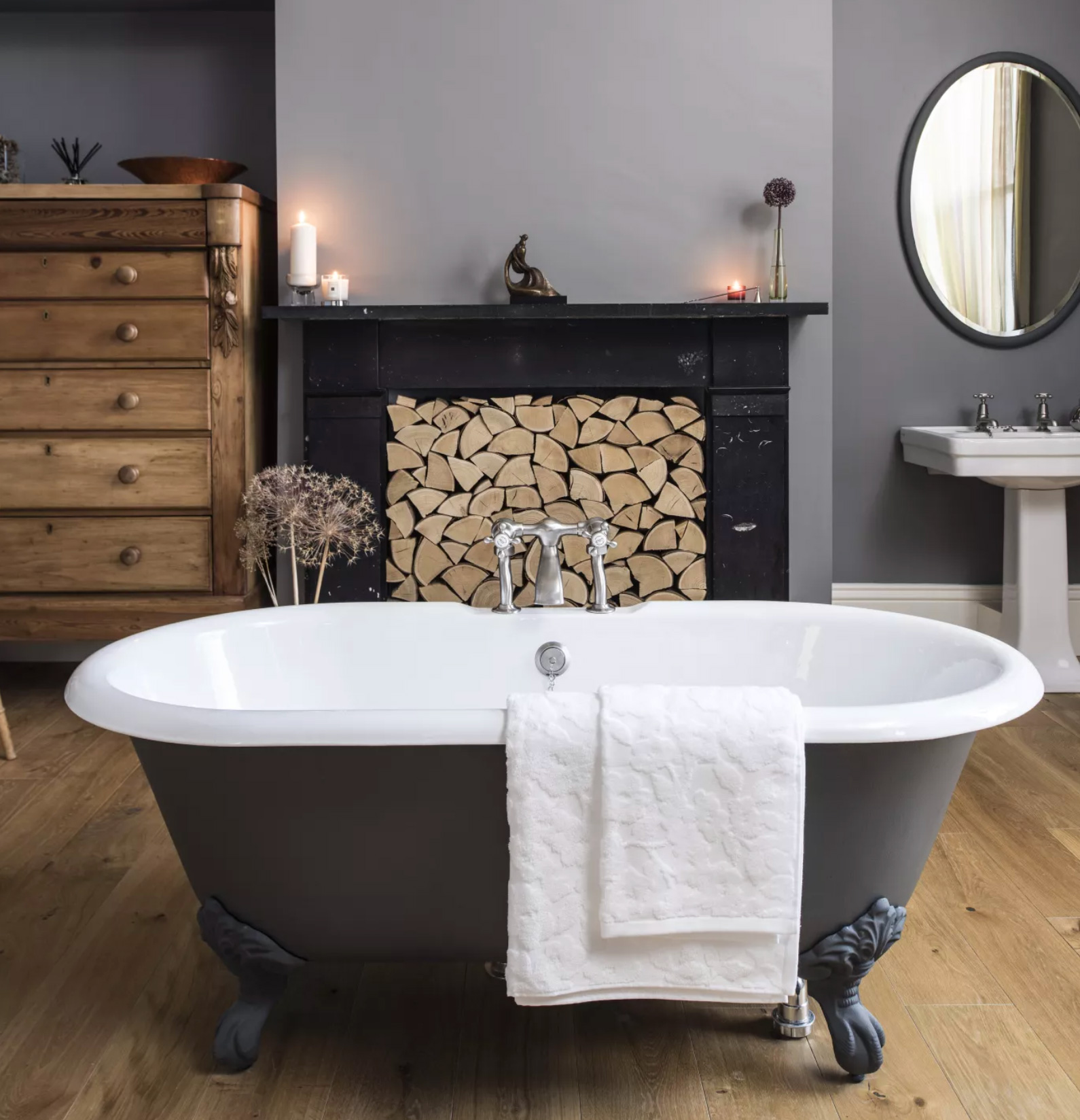 A grey bathroom with a dark grey painted roll top bath and a fireplace filled with logs behind