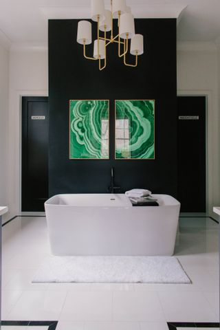 A rectangular white bath tub in a bathroom with black walls and white floor tiles, modern green artwork on the wall and a gold and white pendant light