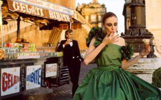 a model in a green dress eating ice cream in the foreground with another model wearing a suit leaning against a ice cream stand in the background.