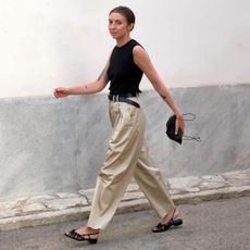 British influencer Brittany Bathgate walking on a street in Molico, Spain wearing a black crewneck tank top, black leather belt, minim black pouch clutch bag, pleated khaki pants, and black woven slingback sandals