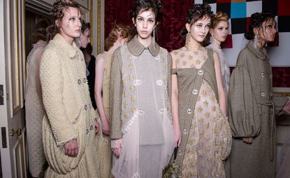 Eight female models wearing ivory, cream and brown embellished and patterned pieces from Simone Rocha's collection. In the background there is a deep red patterned wall and a white and gold door and frame