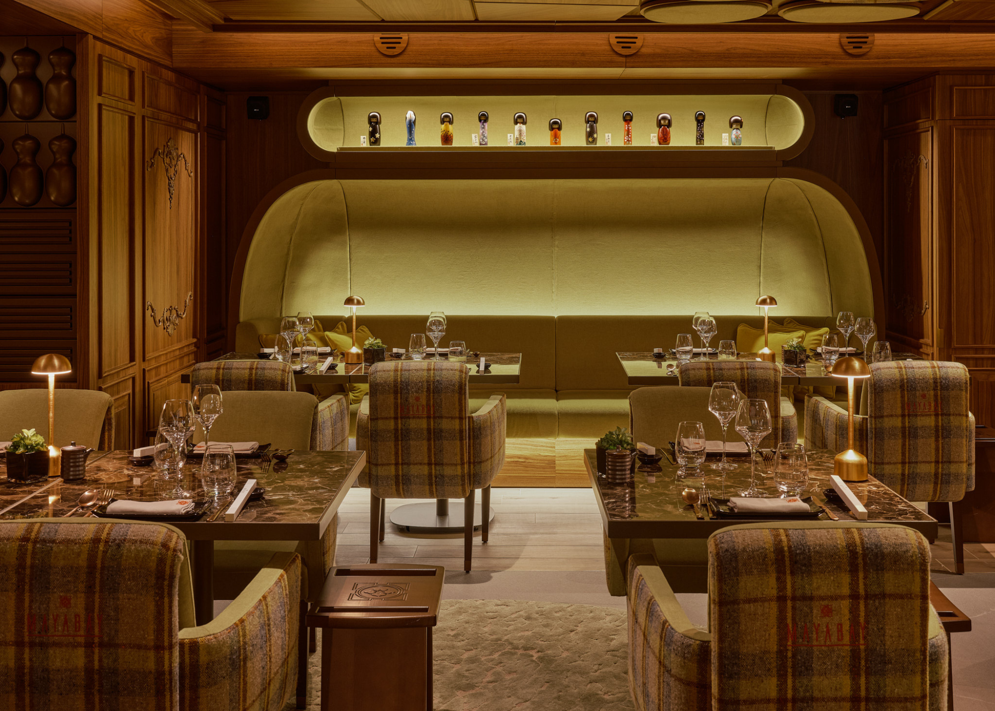 The MayaBay Courchevel restaurant with a space age meets traditional scheme with tartan armchairs at tables with table lamps and a large backlot green banquette seating area at the back with a backlot alcove above with decorative items