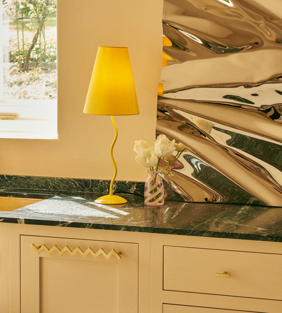 Image of a yellow wavy lamp on a green marble countertop. The cabinets are painted a butter yellow, and there is a gold zigzag pull on the cabinet. On the counter is a large chrome art piece.