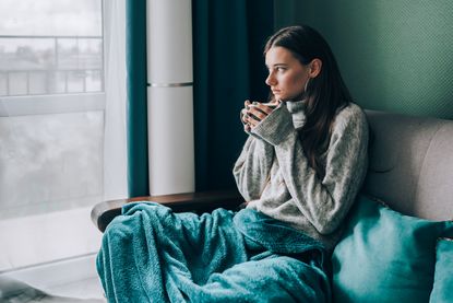 woman in sweater, wrapped in green blue duvet sitting on sofa and drink hot tea