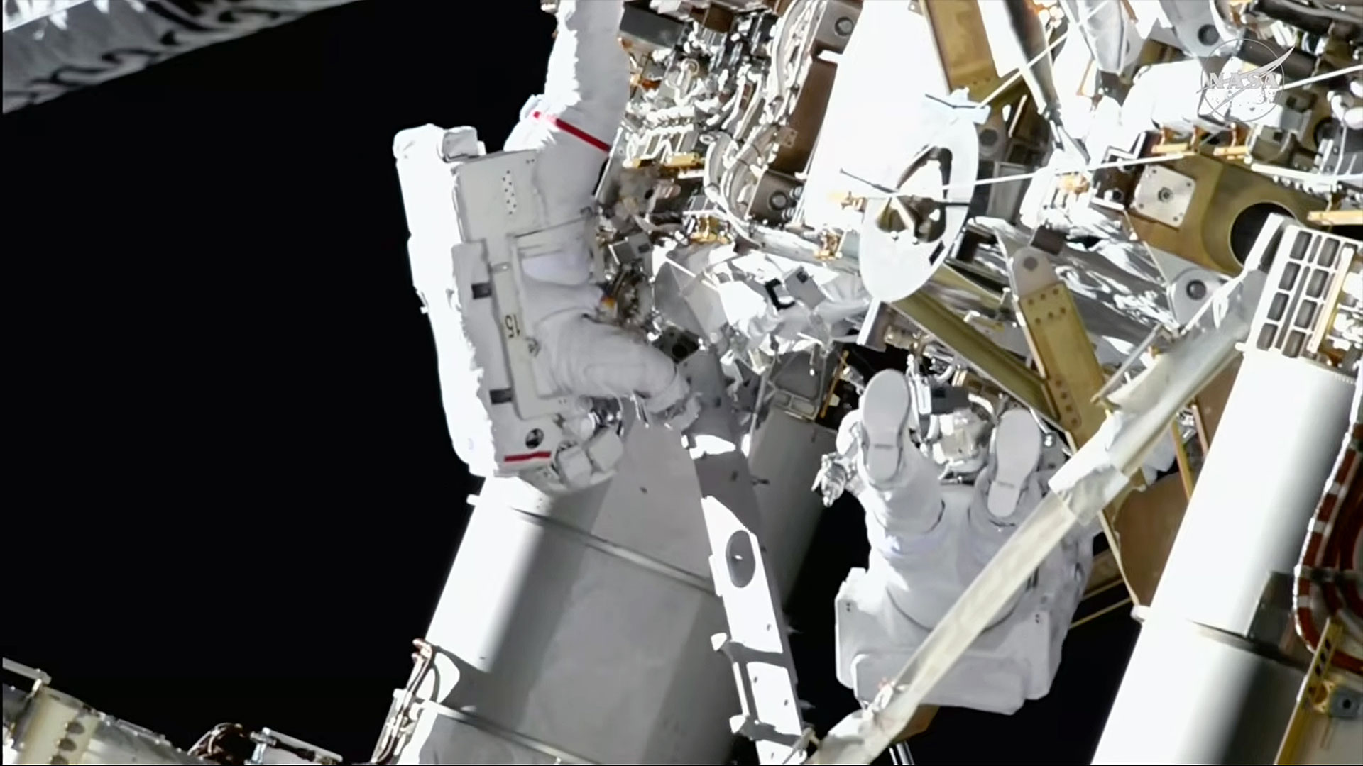 Two astronauts in bulky white spacesuits work outside a large space station, backdropped by the blackness of space.