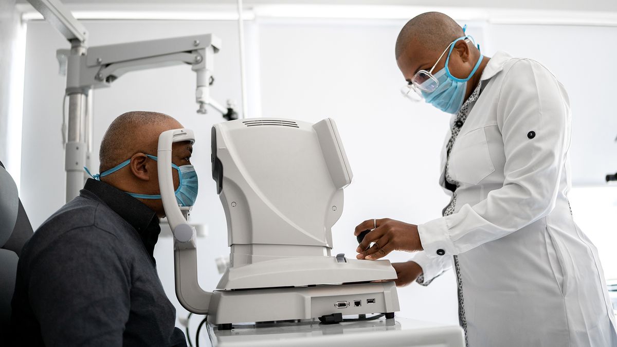 a Black middle-aged man wearing a surgical mask sits in an examination chair in front of an optometry device as a doctor examines his eyes