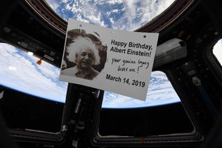 a piece of paper with Albert Einstein's image and the happy birthday, ablert einstein! your genius legacy lives on! March 14, 2019. The paper floats near a window viewing earth from the space station.