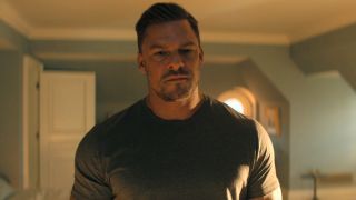 Alan Ritchson as Reacher standing in a bedroom looking down seriously.