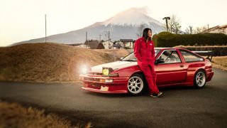 Actor Sung Kang wears a red jumpsuit and sits in front of a red car in in Japan in this promotional image for the Forza Horizon 6 Japan sweepstakes.