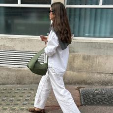 Anna wears a white button down shirt, white jeans, a green bag, leopard-print ballet flats, and a sweater over her shoulders