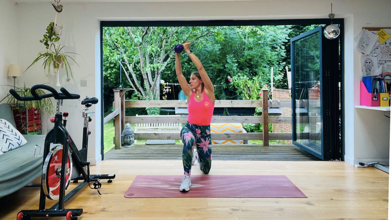 Personal trainer Maddy Biddulph performs a kneeling woodchop. She is wearing a pink vest, tropical print leggings and white sneakers. She is half kneeling, with her left knee on the floor and her right leg extended at a 90 degree angle in front of her. She holds a kettlebell in both hands, up above her head, and her torso is twisting slightly to face her right knee. Besides her is an exercise bike and behind her is a leafy garden.