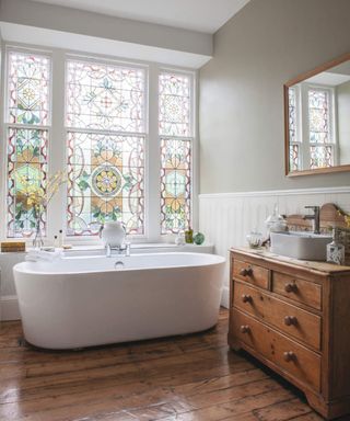 A bathroom with wooden flooring, small white bathtub and stain glassed windows