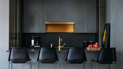 A kitchen in a dark color with a view of the sink and extractor fan as well as bar stools beside the countertop and kitchen area. There is also a few of a chopping board with apples on top of it