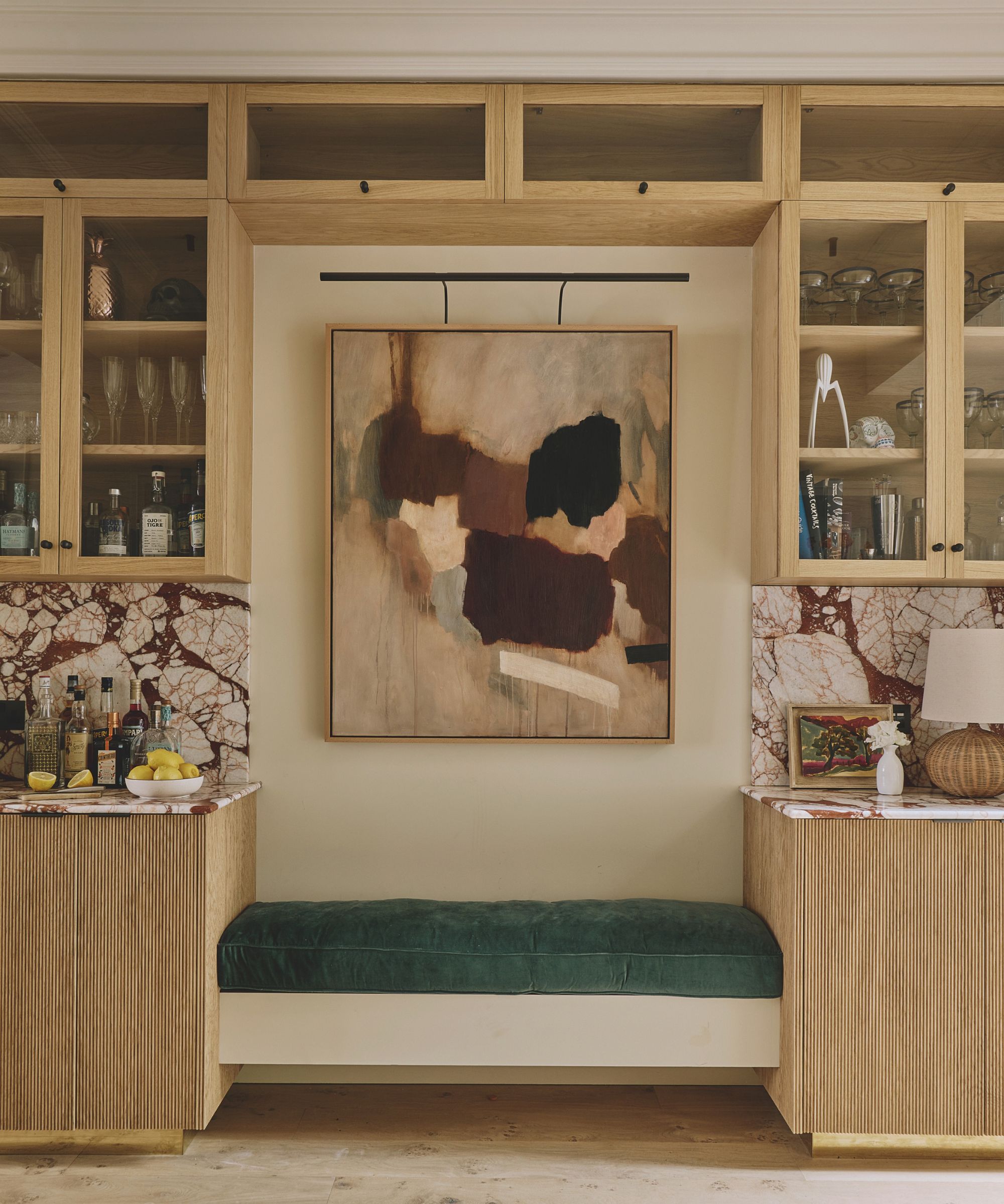 Built-in home bar with oak glass-front cabinets, veined red marble countertops, a green velvet bench seat, and large abstract art framed in the center