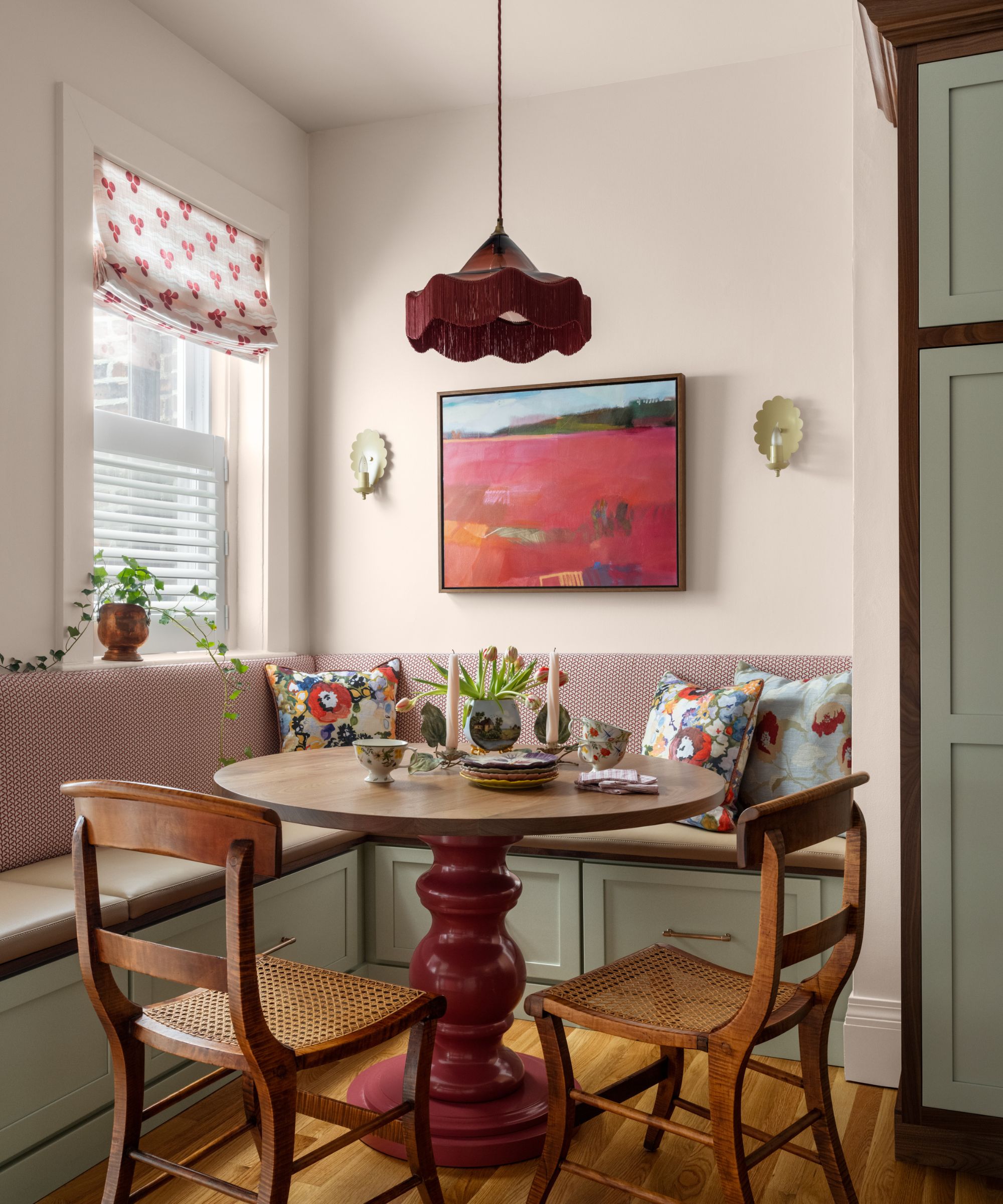 A modern kitchen with a dining nook with white walls, banquette seating with red striped upholstery. A round table with a glossy red base, two wood chairs, and a pink landscape artwork on the wall.