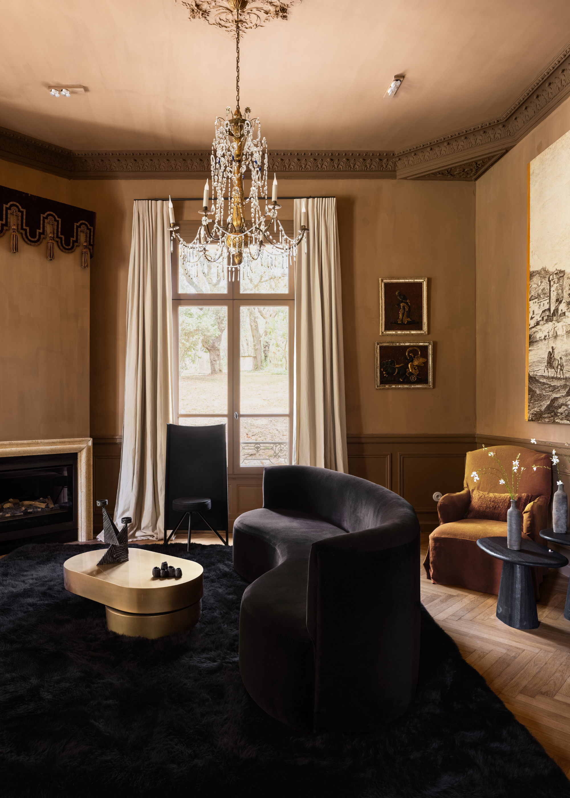 A maximalist living room with a hanging chandelier, a curved soft dark blue sofa, a brushed gold coffee table, and an ochre armchair