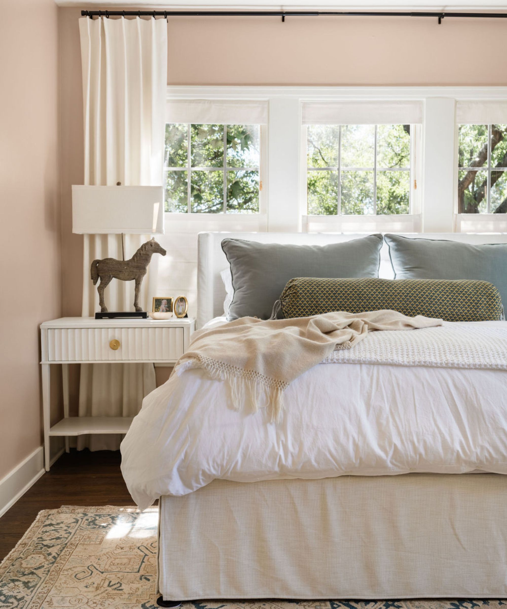 A light and airy bedroom with light pink walls, white bedding, and three windows above the headboard.
