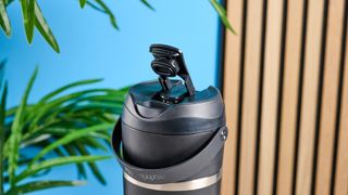 the Nutribullet Flip with a black insulated tumbler and a portable battery-powered blade photographed with a blue background. The tumbler has a carry strap and a sippy lid you can interchange, and an on/off button on the base.