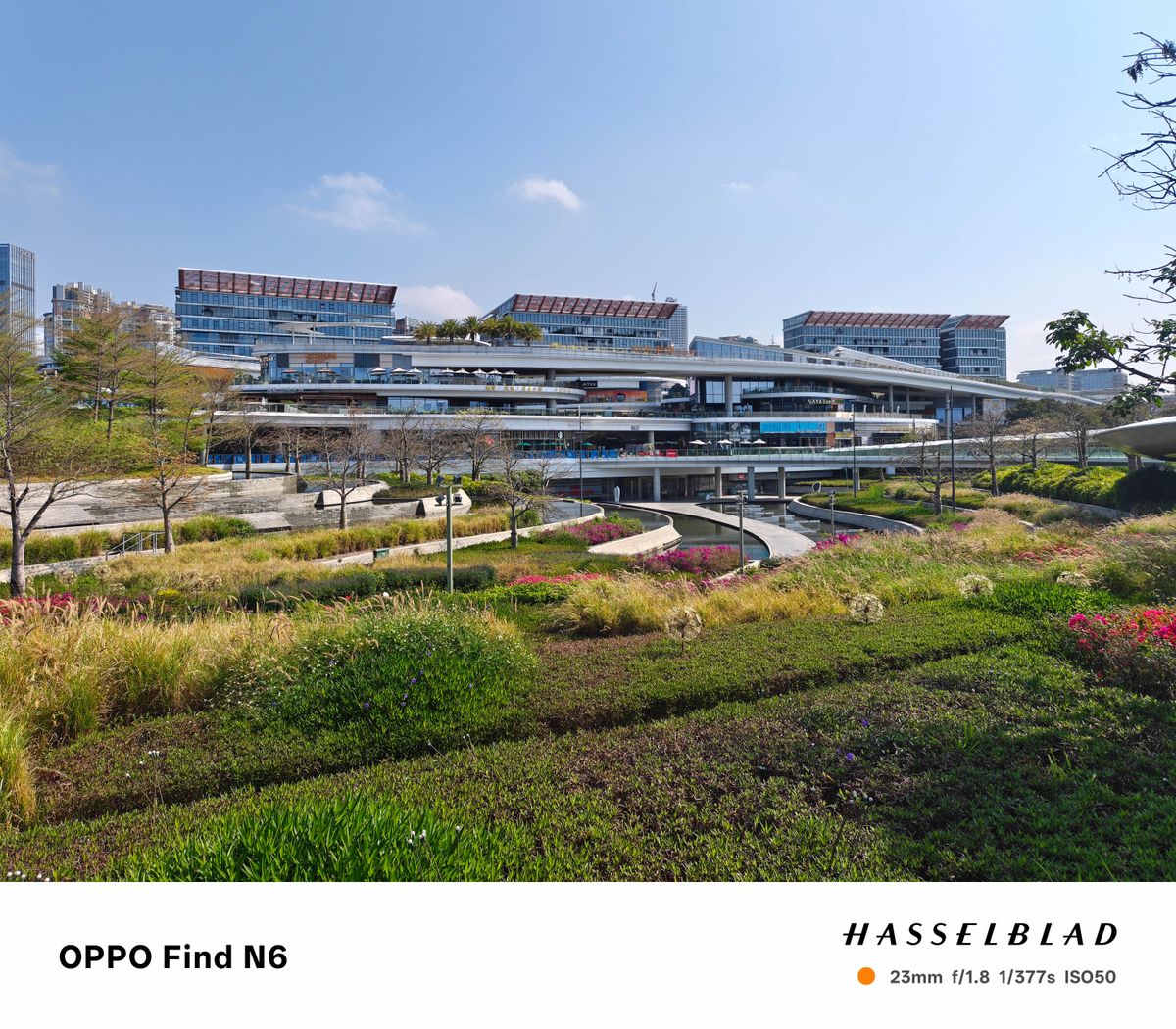 A wide-angle shot of a contemporary architectural development featuring several levels of shops and restaurants. The foreground is filled with a manicured garden of green shrubbery and tall grasses, while large glass-walled buildings rise in the background.