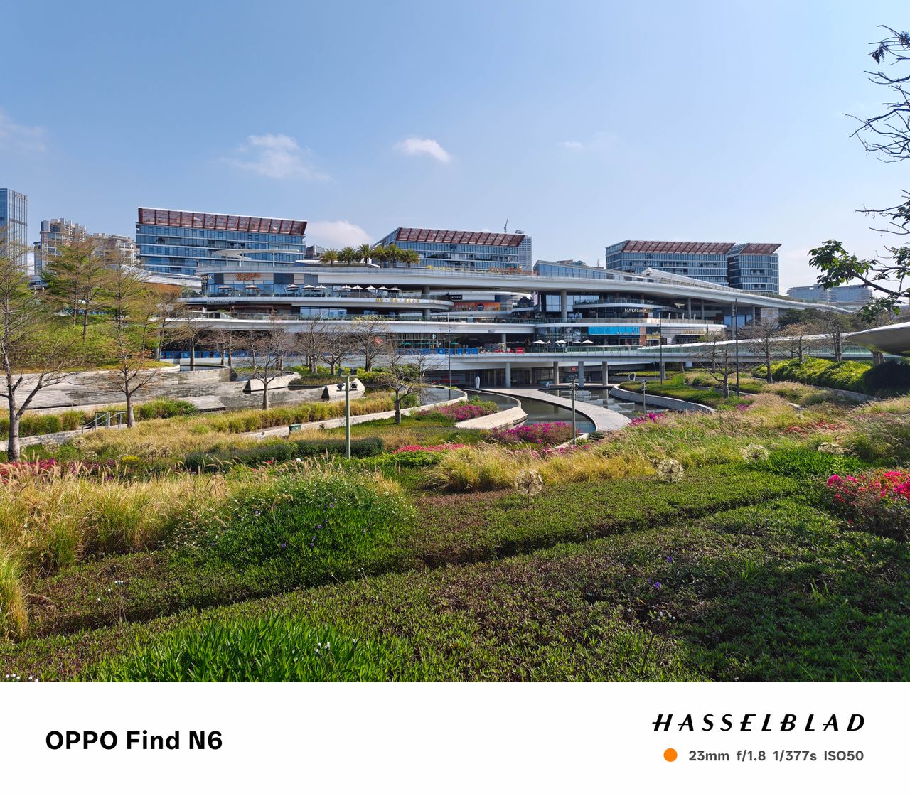 A wide-angle shot of a contemporary architectural development featuring several levels of shops and restaurants. The foreground is filled with a manicured garden of green shrubbery and tall grasses, while large glass-walled buildings rise in the background.