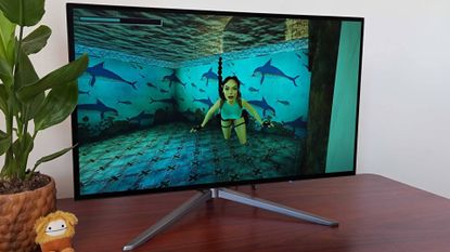 KTC G27P6 OLED gaming monitor on woodgrain desk next to plant with Lara Croft swimming on screen.