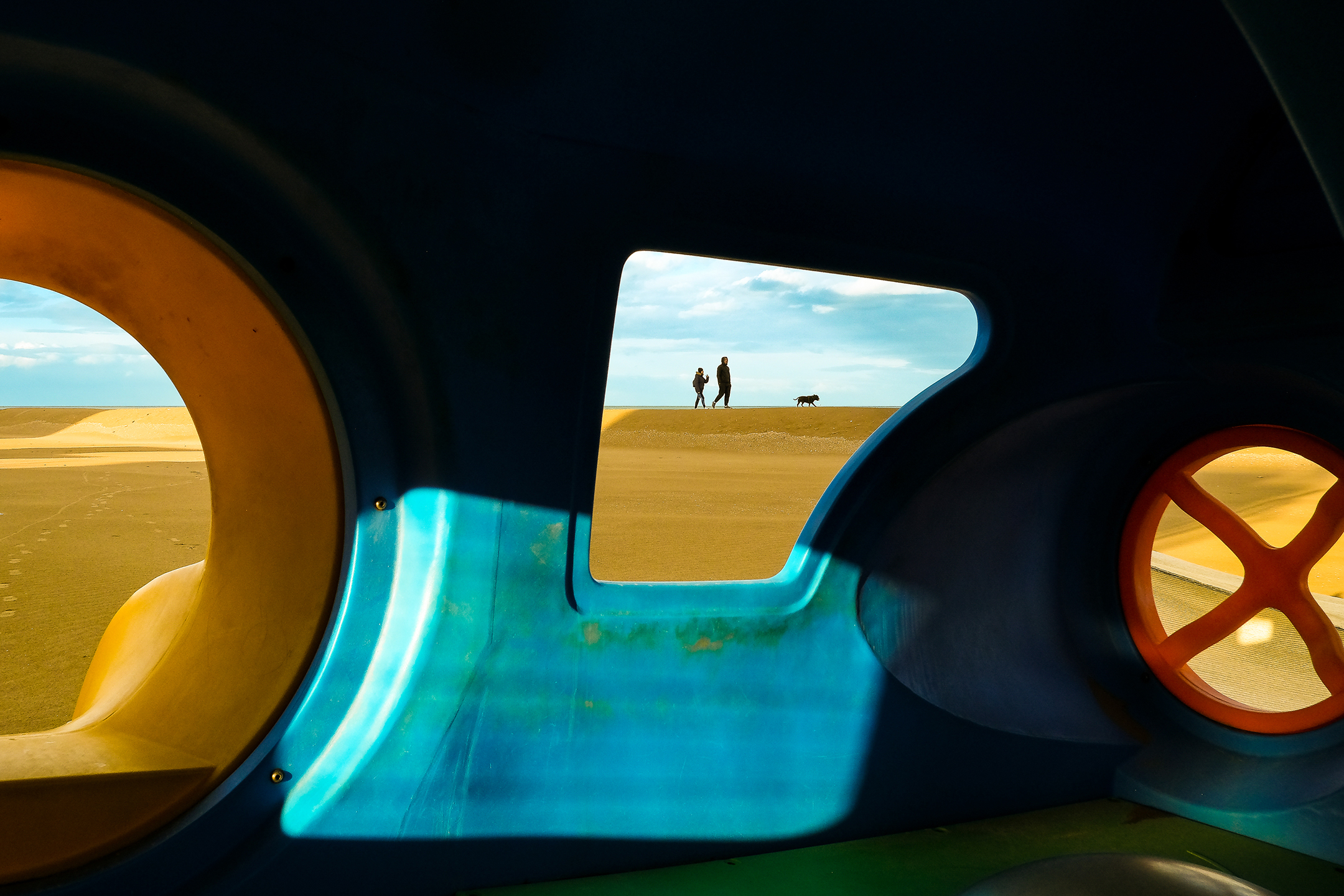 View through colorful playground tunnel framing a couple with a dog on dunes under a blue sky. Bright, playful, and serene atmosphere