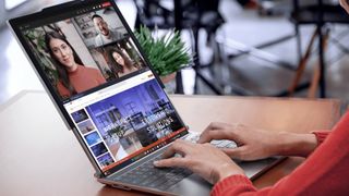 A woman is typing at a Lenovo ThinkBook Plus Gen 6 Rollable laptop in a well-lit office. The screen is angled towards our view and a small potted plant is seen poking out from behind the laptop. The screen is fully extended, rocking a tall 8:9 aspect ratio. The top portion of the screen is hosting a video call with three particpants, while the bottom, extended portion of the screen shows the woman typing is working on a slideshow deck about ergonomic workspace solutions.