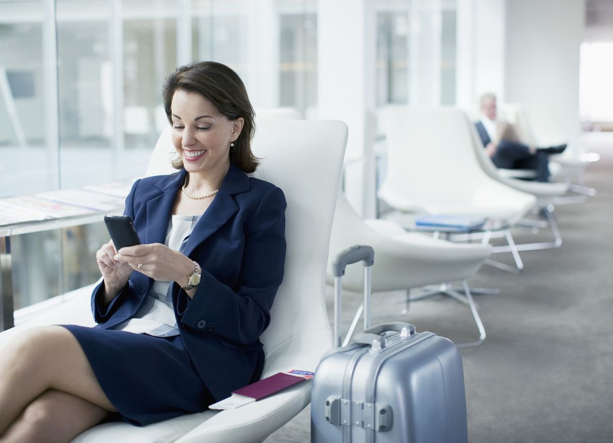 Businesswoman with luggage sitting in airport waiting area smiling at phone