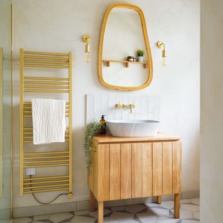 a small neutral bathroom with a pale wood vanity unit, matching framed mirror and gold coloured towel rail