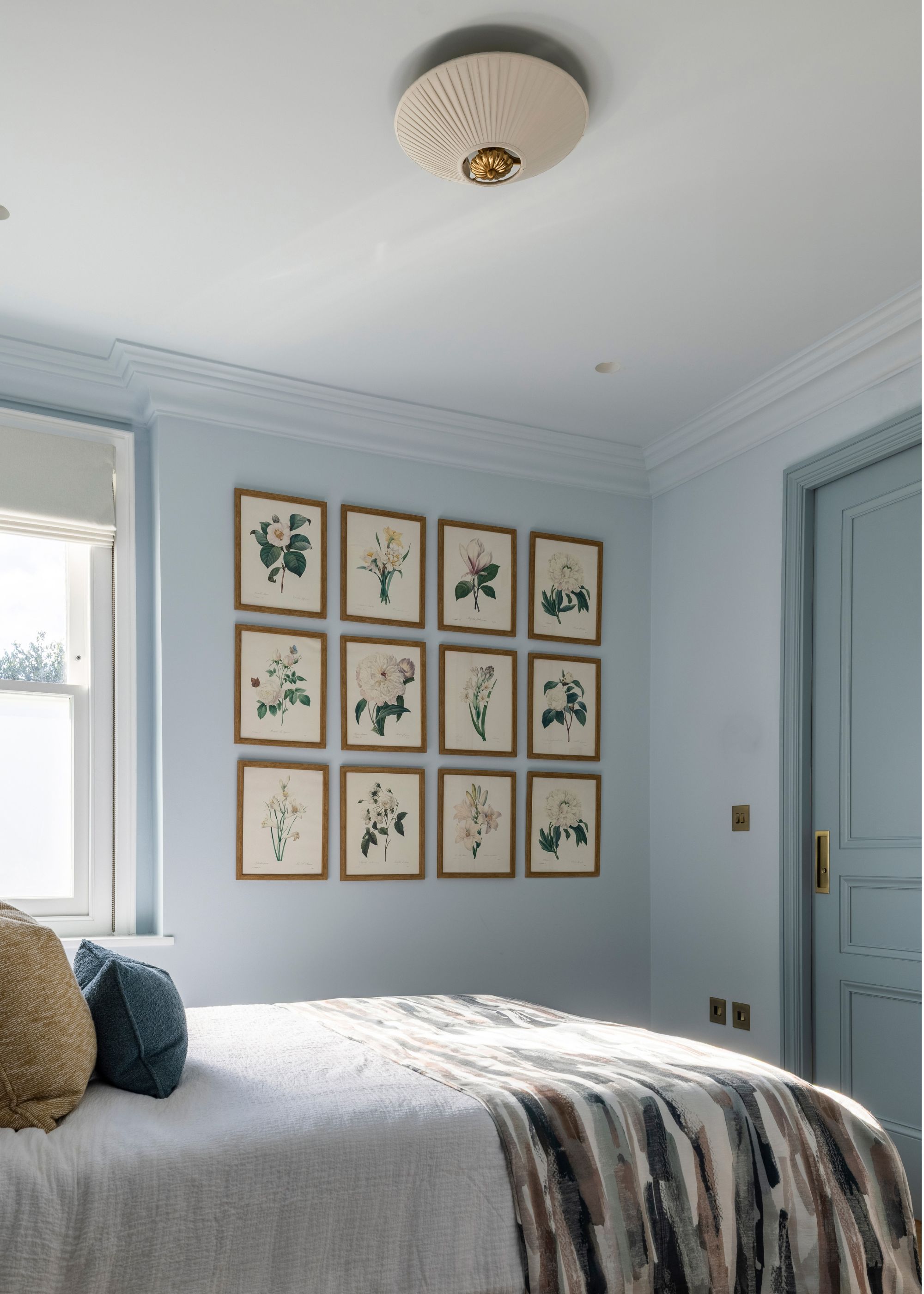 A bedroom with pale blue walls and darker blue paint on the door and woodwork. A bed with layered neutral bedding, and a gallery wall of botanical prints.