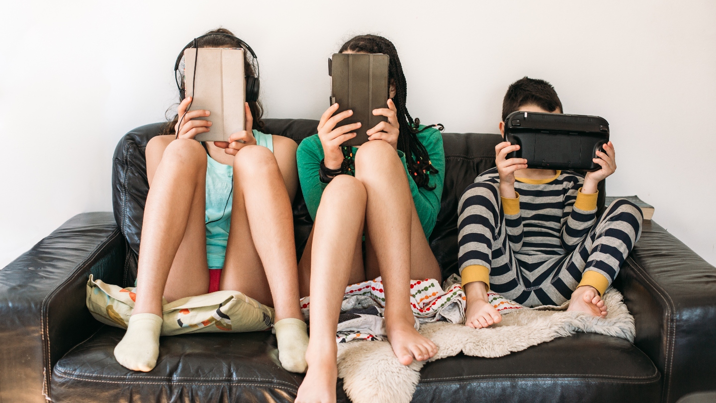 Three kids sitting on a couch using technology and holding the devices in front of their faces