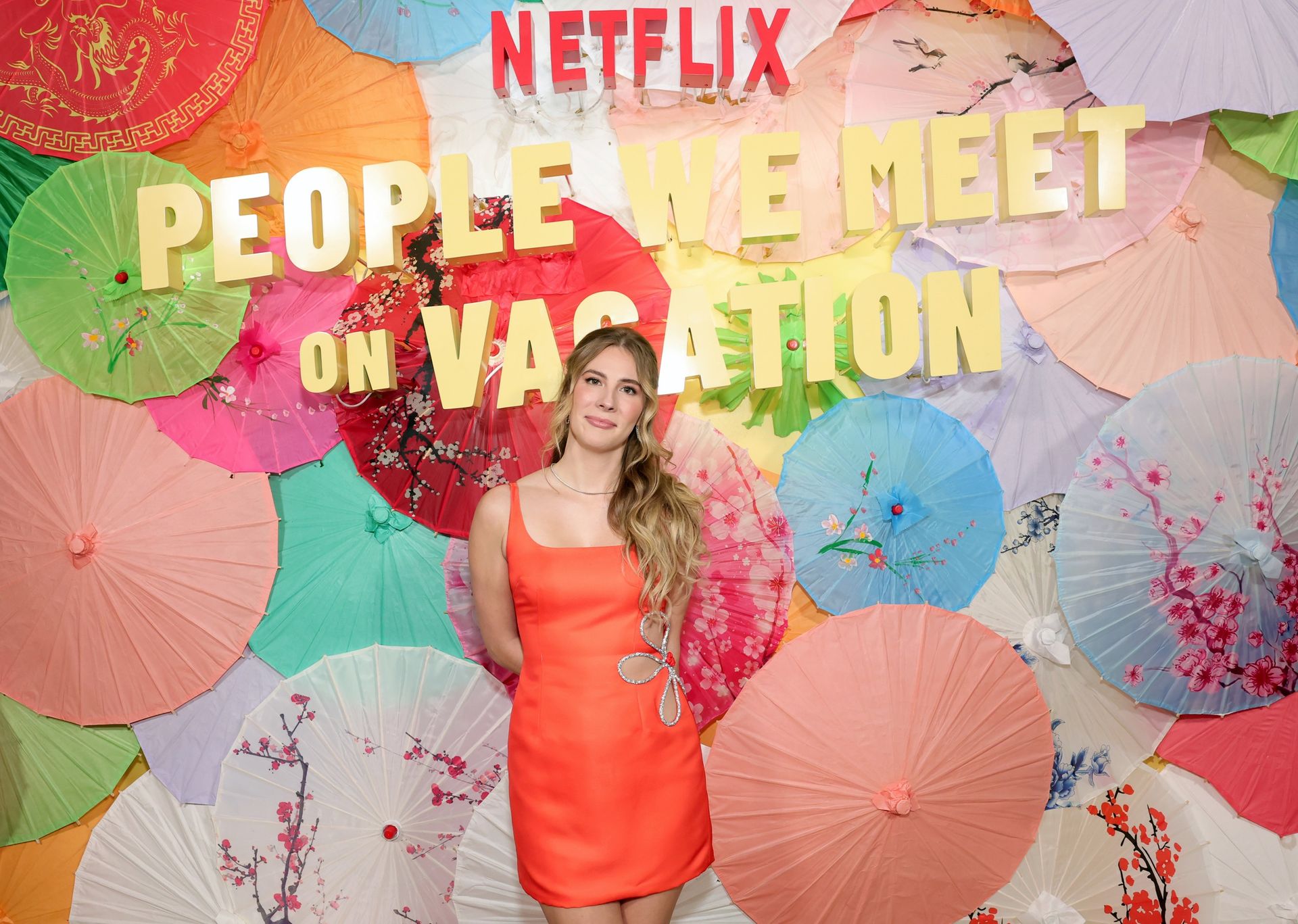 emily henry wears an orange dress and poses in front of a wall of umbrellas at a premiere event for people we meet on vacation