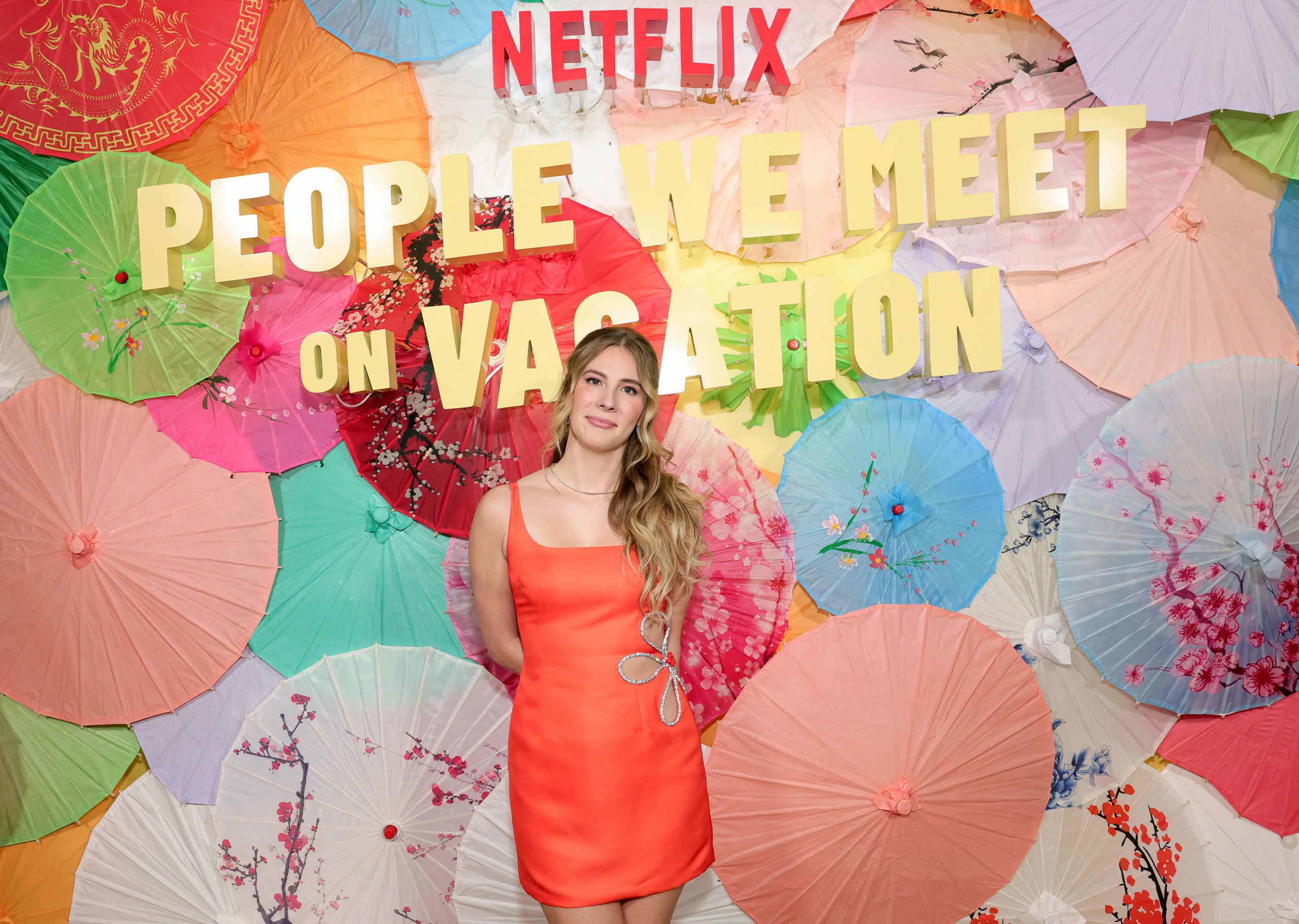 emily henry wears an orange dress and poses in front of a wall of umbrellas at a premiere event for people we meet on vacation