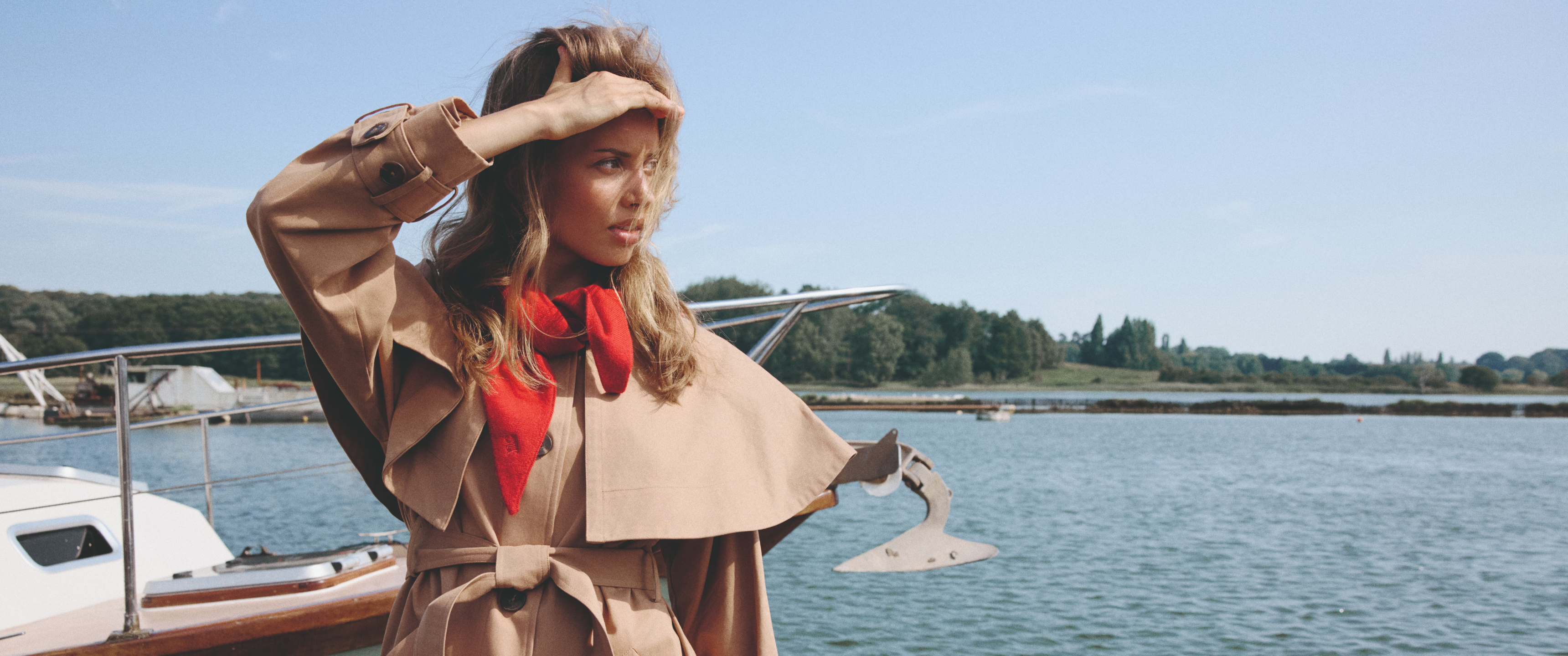 Model by a boat wearing a tan trench coat and a red scarf sold at nobody's child