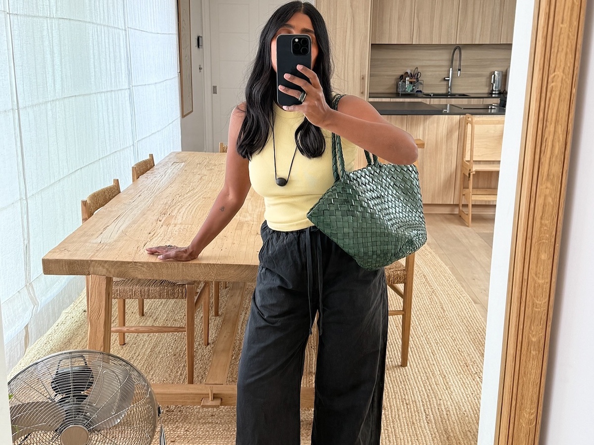 British influencer Monikh Dale poses for a mirror selfie in her London living room with a wood table, rattan chairs, and jute rug while wearing a butter-yellow tank top, woven green tote bag, cord necklace with a black pendant, and black linen pants.