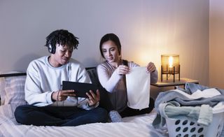 couple sitting on bed at night - one is folding laundry and the other is using a tablet