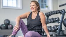 A smiling woman in leggings and a tank top sits in a gym. Behind her we see a rack of dumbbells and some medicine balls.
