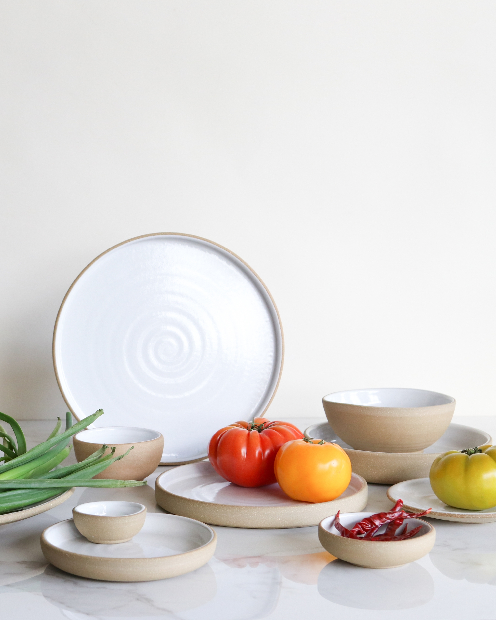 Image of a set of white ceramic plates and bowls arranged with tomatoes and chilis on them in front of a white background and marble countertop.