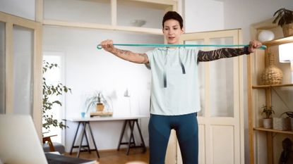 woman standing facing the camera and a laptop holding a turquoise resistance band in front of her chest pulling it wide apart. she's in a home setting with double doors and a bookcase behind her.