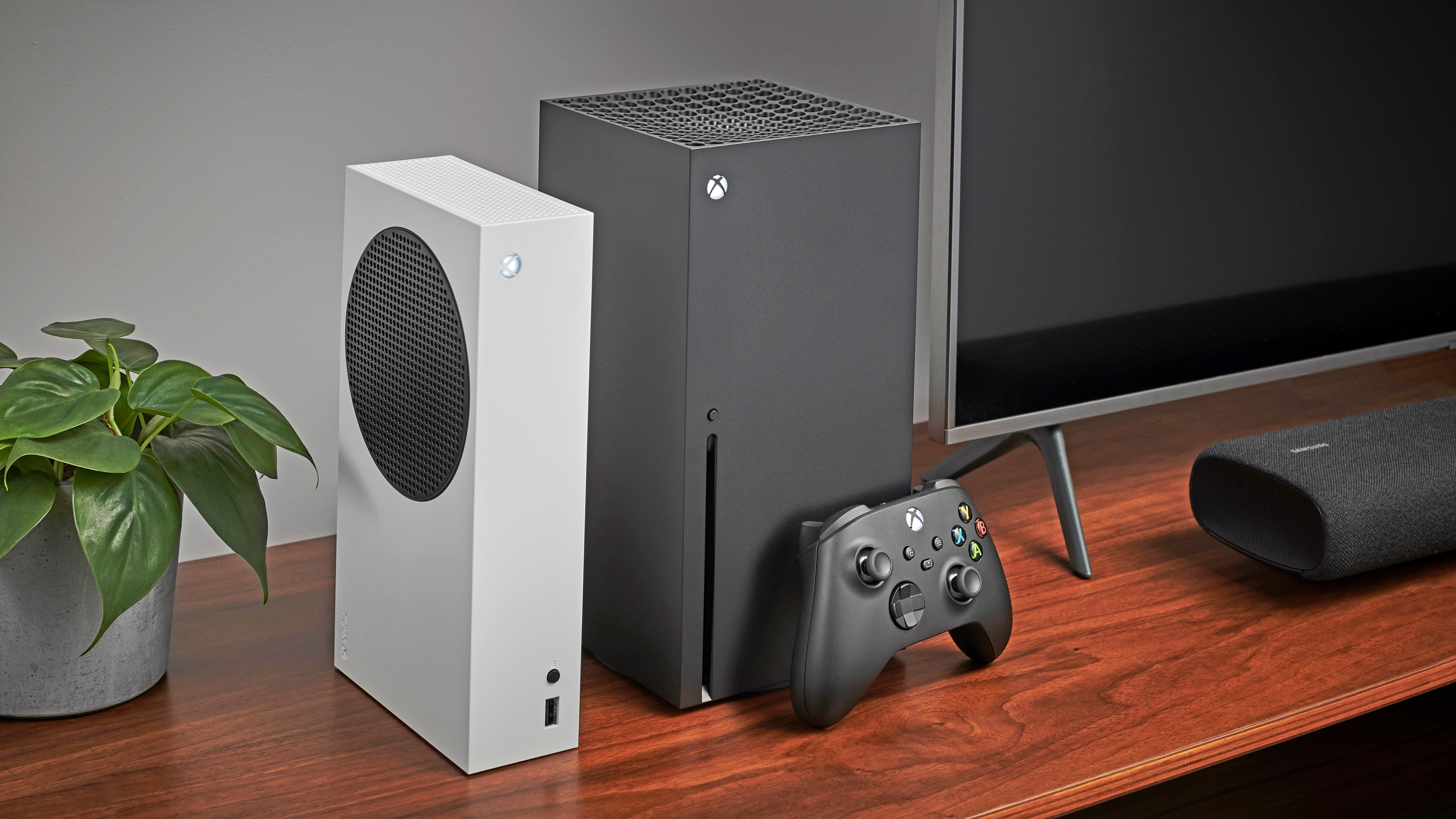 An image of an Xbox Series X next to an Xbox Series S on a wooden TV stand next to a TV, with a black controller leaning on the Xbox Series X.