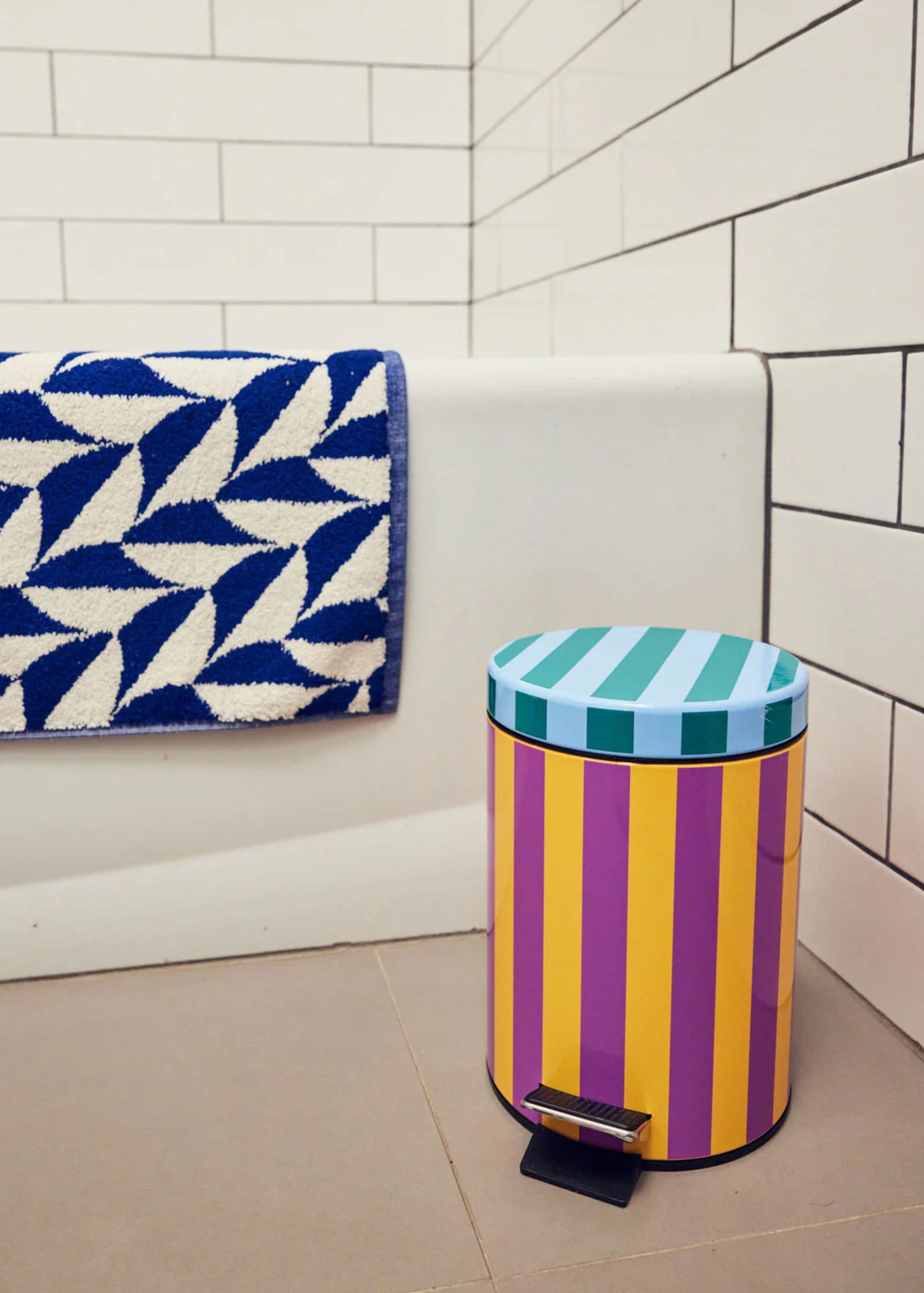 A striped and colorful trash can beside a bathtub that also has a white and blue towel on the side 
